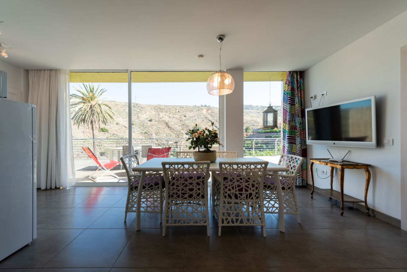 Zona pranzo con tavolo e sedie, televisione a parete e vista su un paesaggio desertico.