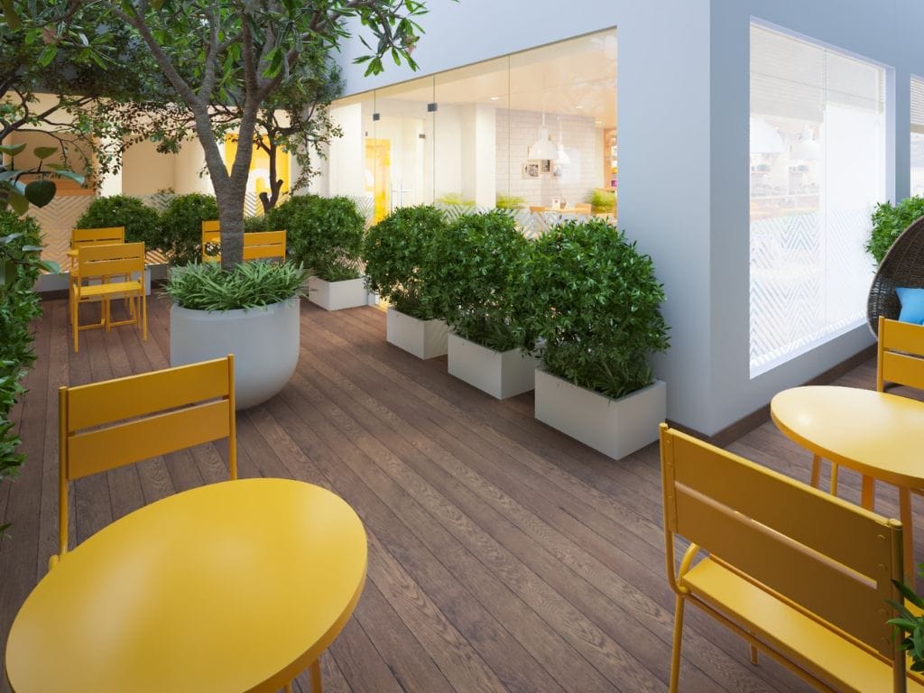 Un espacio interior con asientos, piso de madera, sillas amarillas y macetas blancas con plantas verdes.
