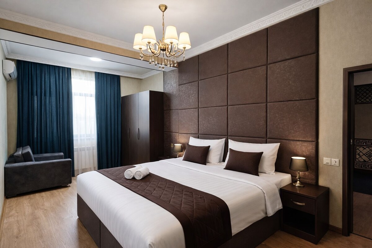 Habitación de hotel con una cama grande, un cabecero oscuro, una araña, una mesita de noche y un área de estar con una silla y cortinas.