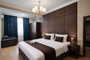 Habitación de hotel moderna con una cama grande, cabecera oscura, candelabro, zona de estar con sofá y vistas al exterior a través de una ventana con cortinas.