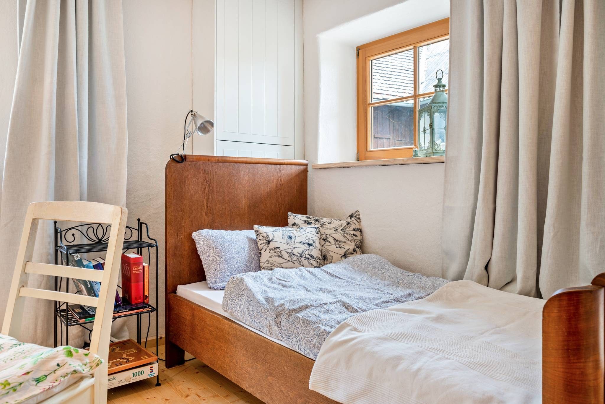 Ein Einzelbett mit Holz-Kopfteil, ein Nachttisch mit Lampe und ein Fenster mit Vorhängen.