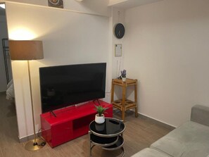 A living room with a flat-screen TV on a red stand, a small round table with a plant, a wooden stool, and a grey sofa.