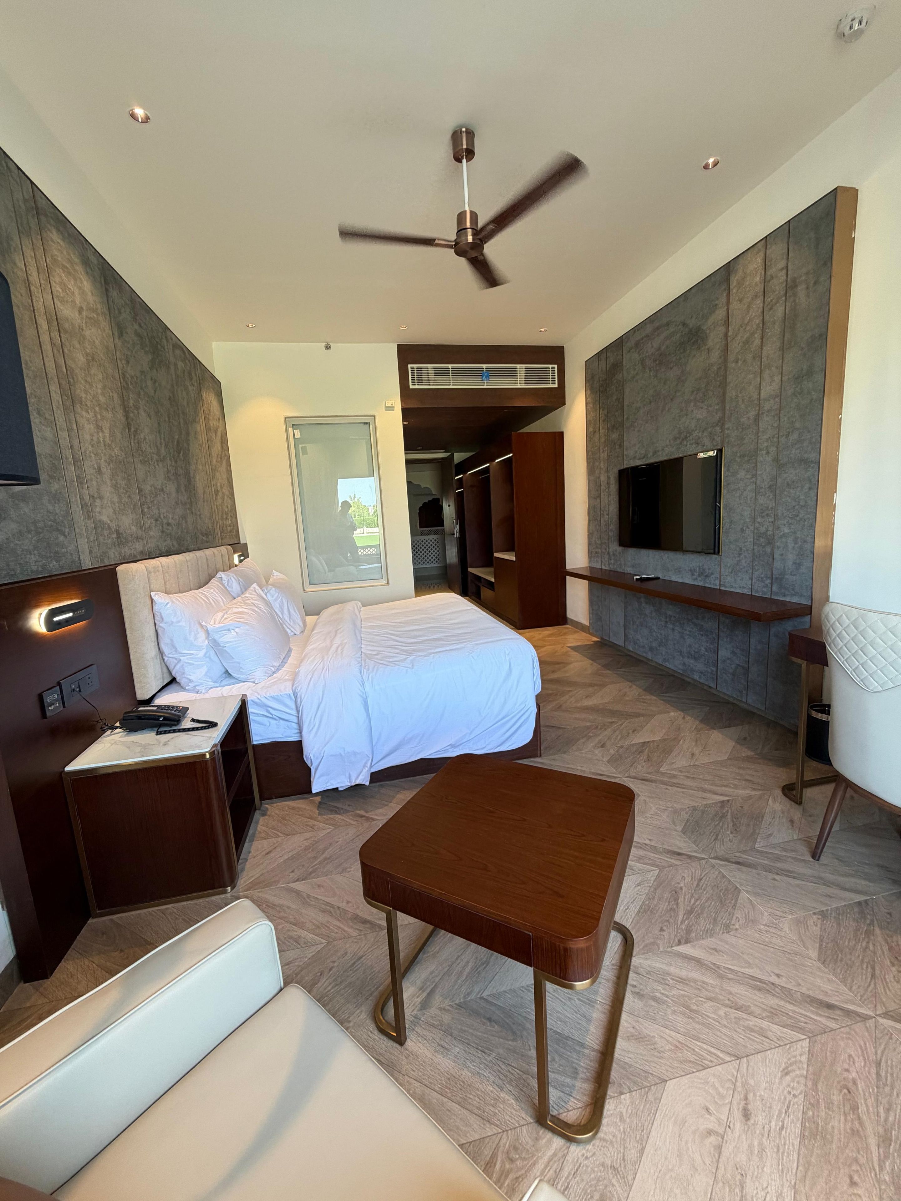 Una habitación de hotel moderna con cama, mesita de noche, un pequeño rincón de descanso, ventilador de techo y televisión montada en la pared.
