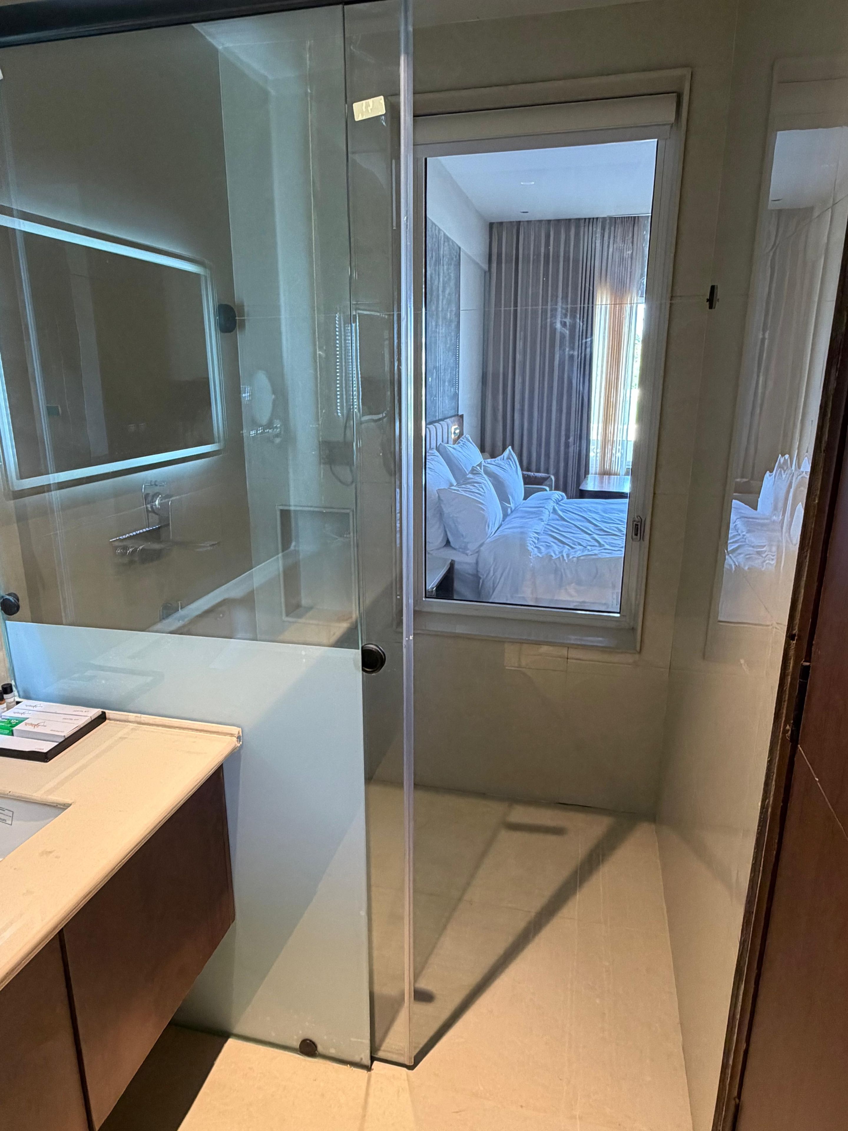 Habitación de hotel con separador de vidrio, cama con ropa blanca, mesita de noche y ventana con cortinas.