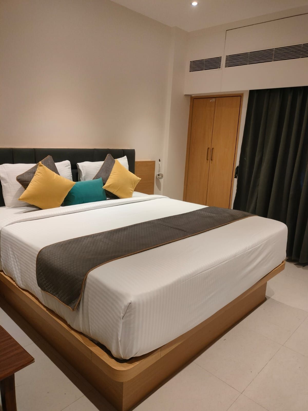 Habitación de hotel con cama, mesita de noche de madera, armario y ventana con cortinas.
