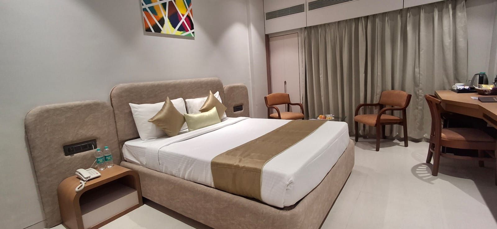 A hotel room with a bed, bedside table, two chairs, a desk, and a large window with curtains.