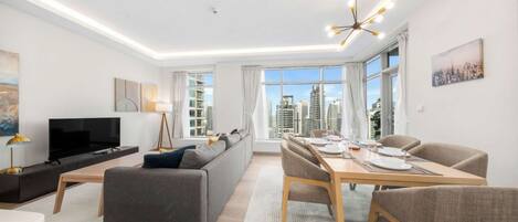 A modern living room with a flat-screen TV, grey sofa, dining table set for four, and large windows offering a city view.