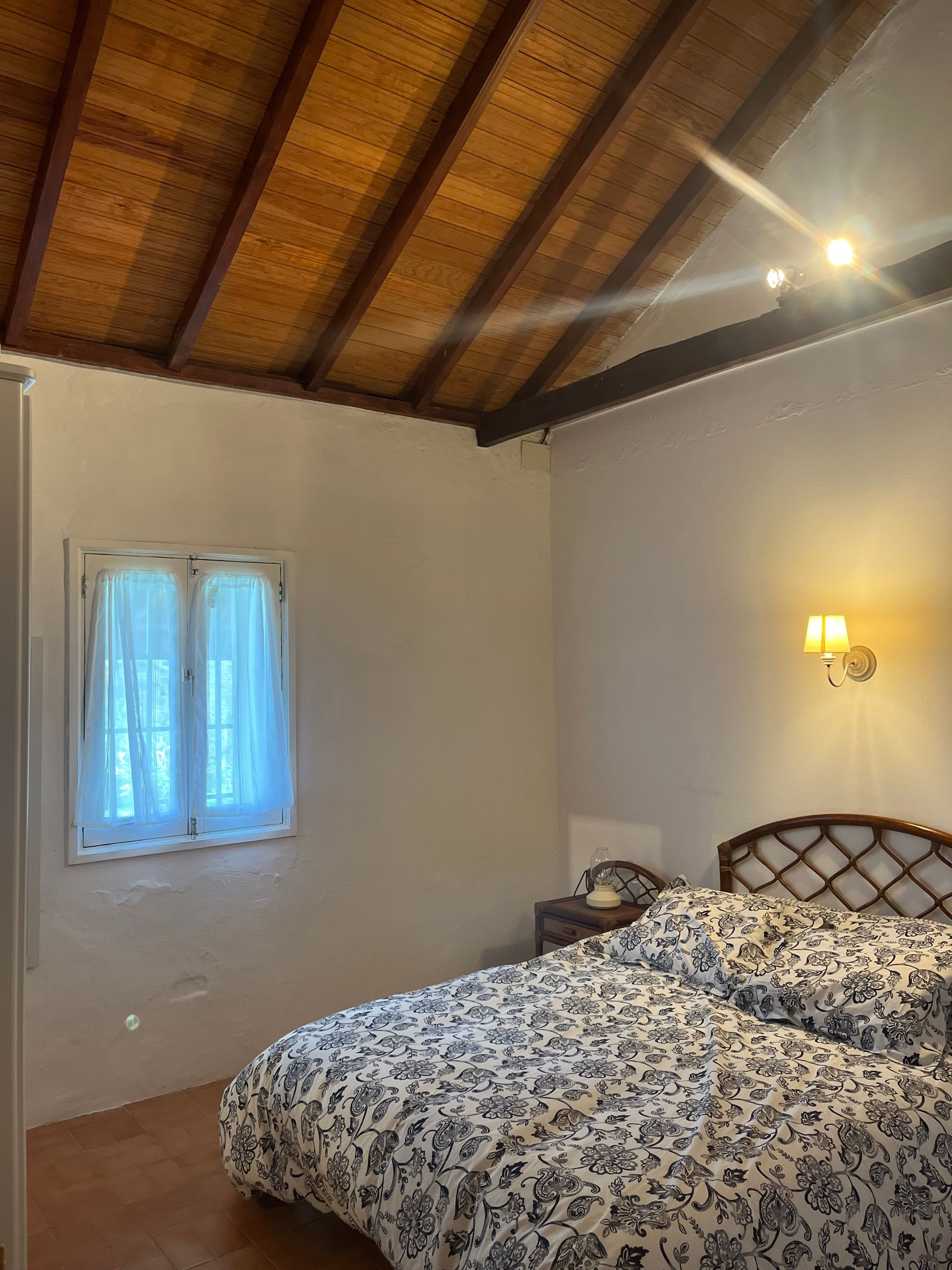Une chambre avec un plafond en bois, un lit recouvert d’une couvre-lit à motifs, une table de chevet avec une lampe et une fenêtre avec des rideaux.