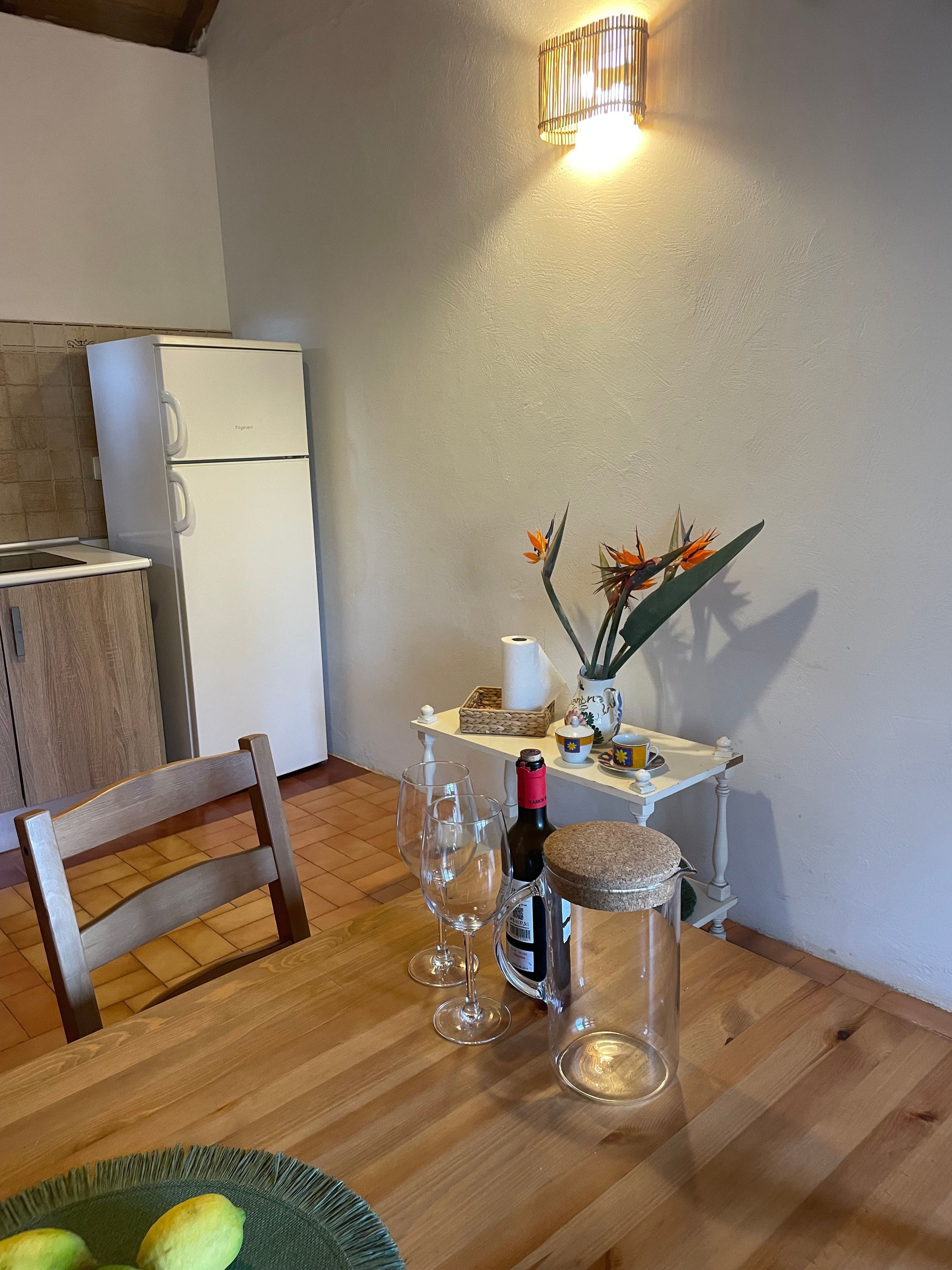 Une petite cuisine avec un réfrigérateur blanc, une table à manger en bois, deux verres à vin, une bouteille de vin et une plante décorative.