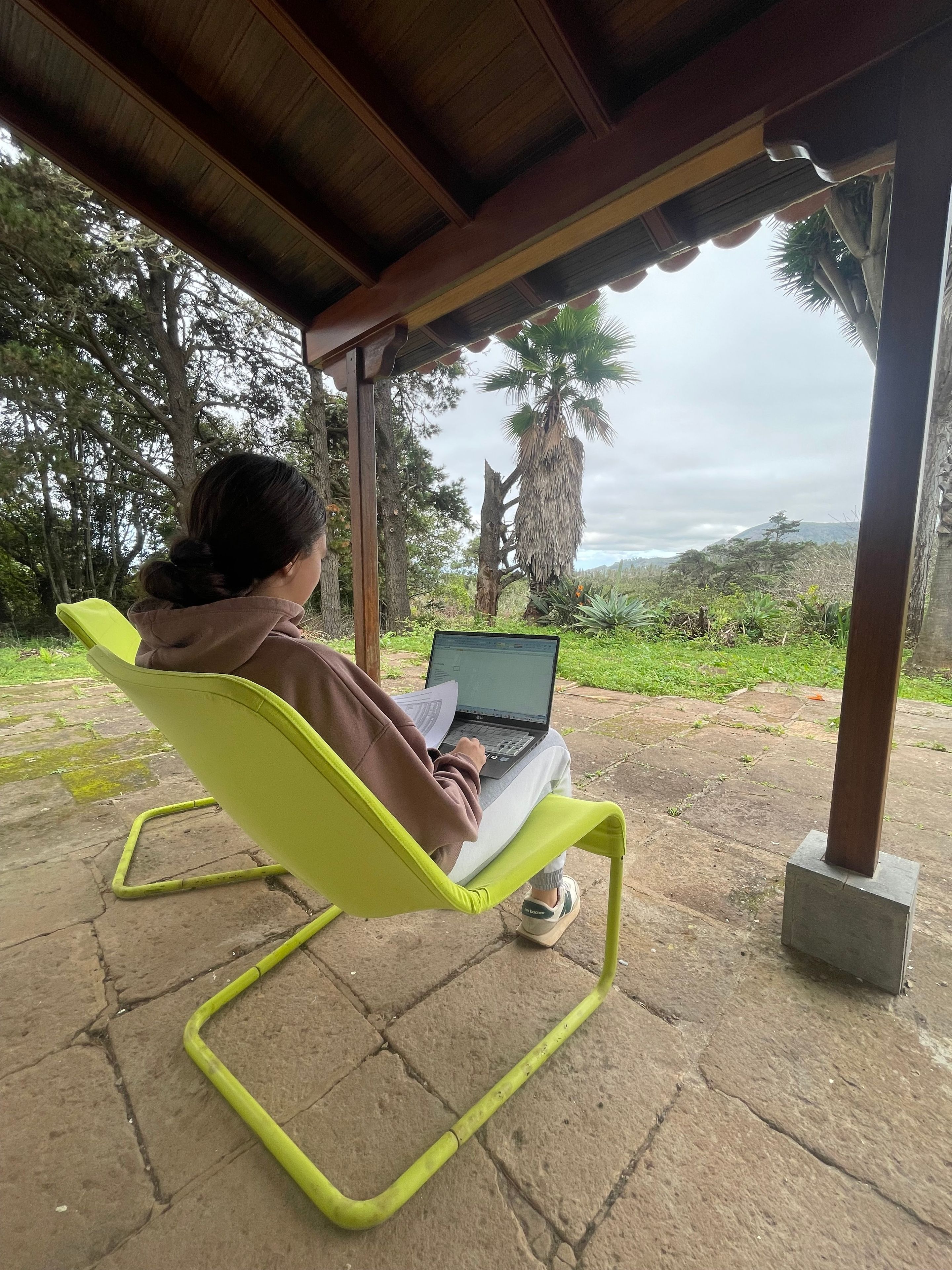 Une personne est assise sur une chaise jaune vif, un ordinateur portable sur les genoux, sous une structure en bois d’où l’on peut admirer des arbres et des collines.