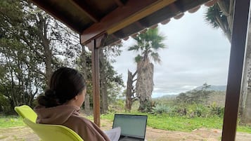 Une personne est assise sur une chaise jaune vif, un ordinateur portable sur les genoux, sous une structure en bois d’où l’on peut admirer des arbres et des collines.