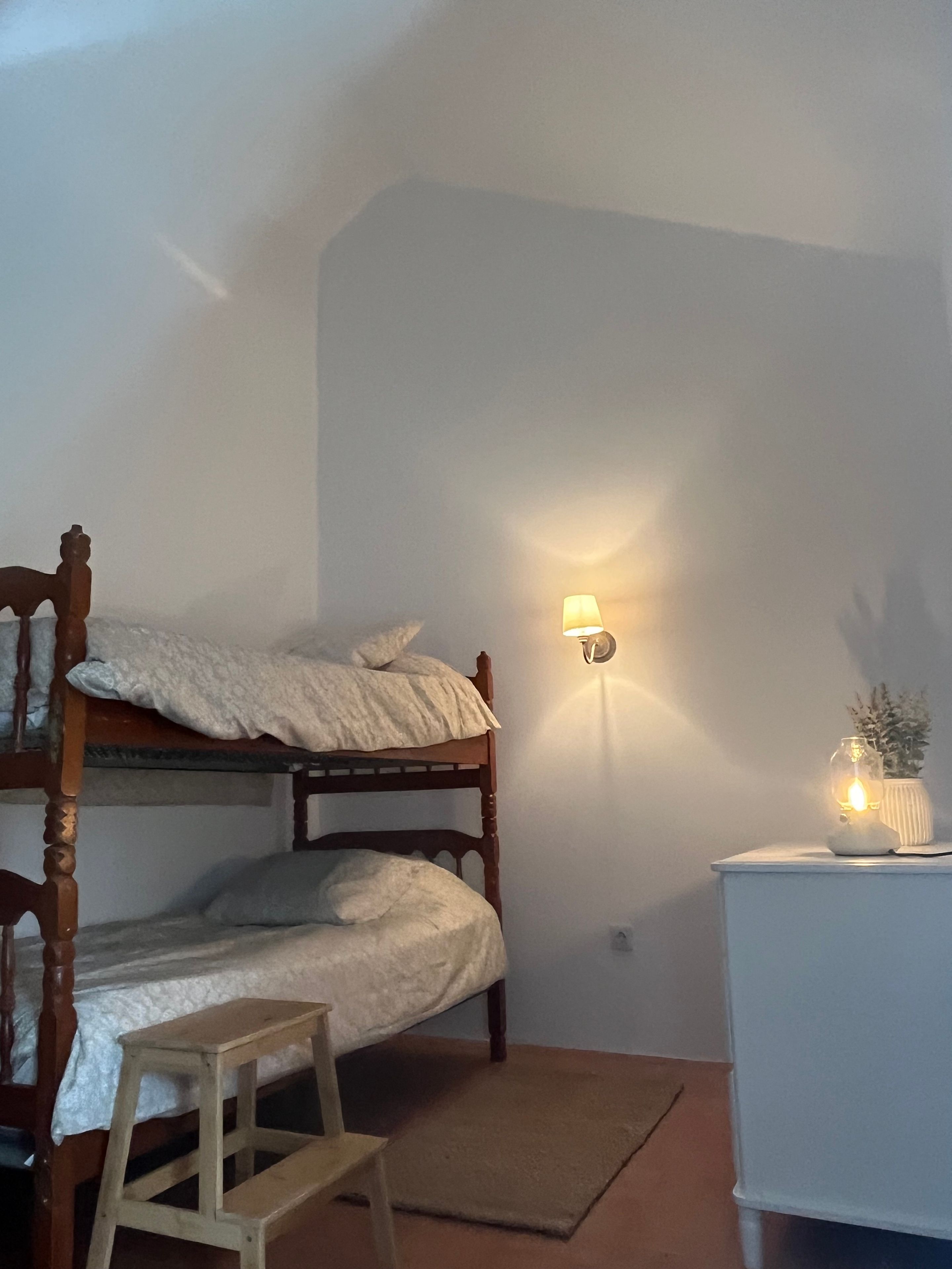 Un lit superposé avec une structure en bois et du linge de lit blanc, un petit tabouret en bois, une table de chevet blanche avec une lampe et une plante en pot.
