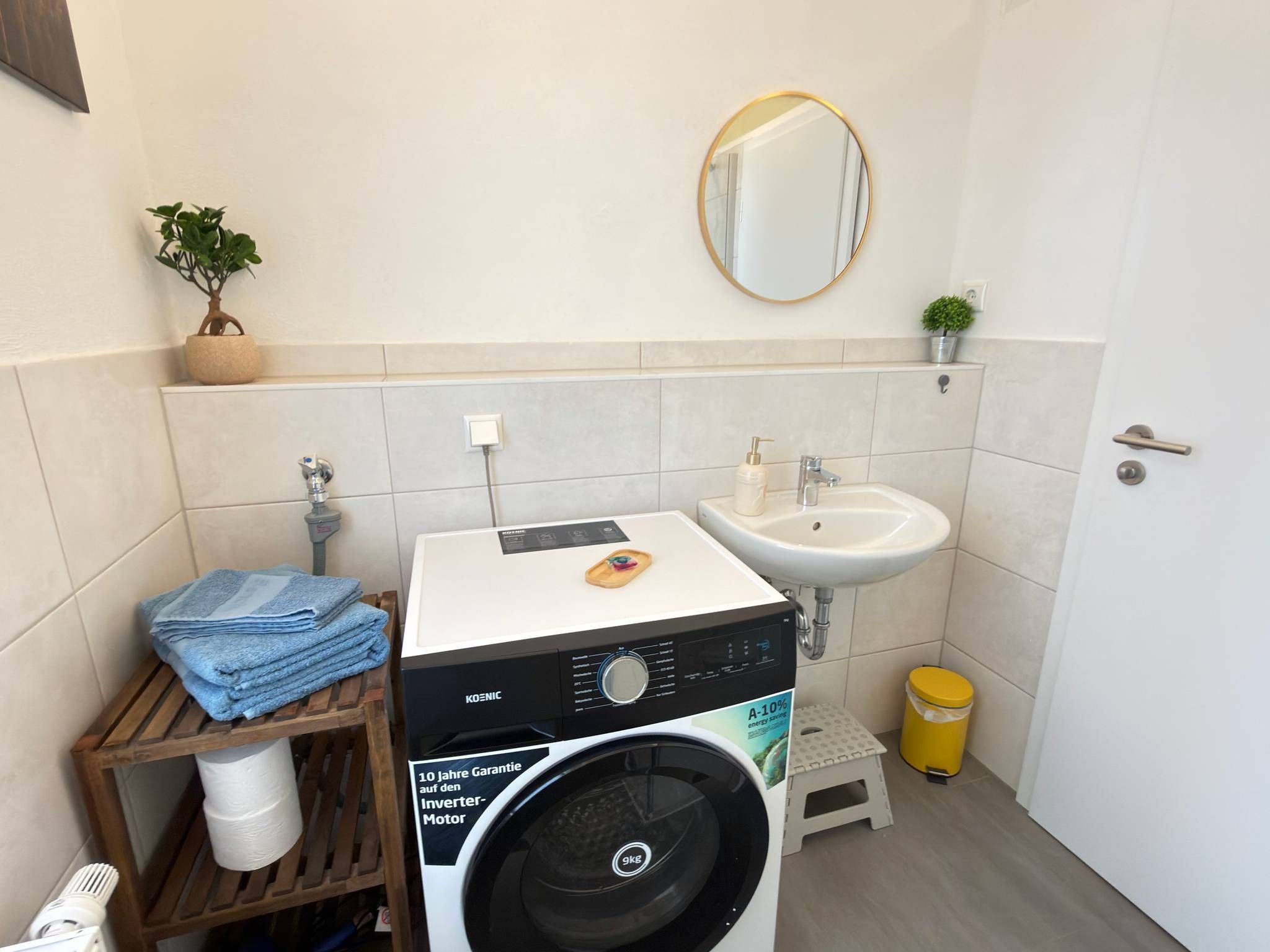 Ein kleines Badezimmer mit Waschmaschine, Waschbecken und einer kleinen Bank.