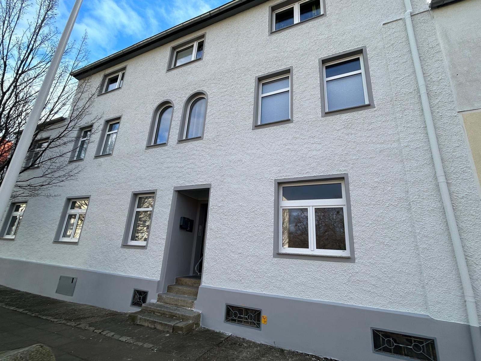 En bygning med flere etager, en hvid facade, mange vinduer og en tydelig indgang med trapper op til døren.