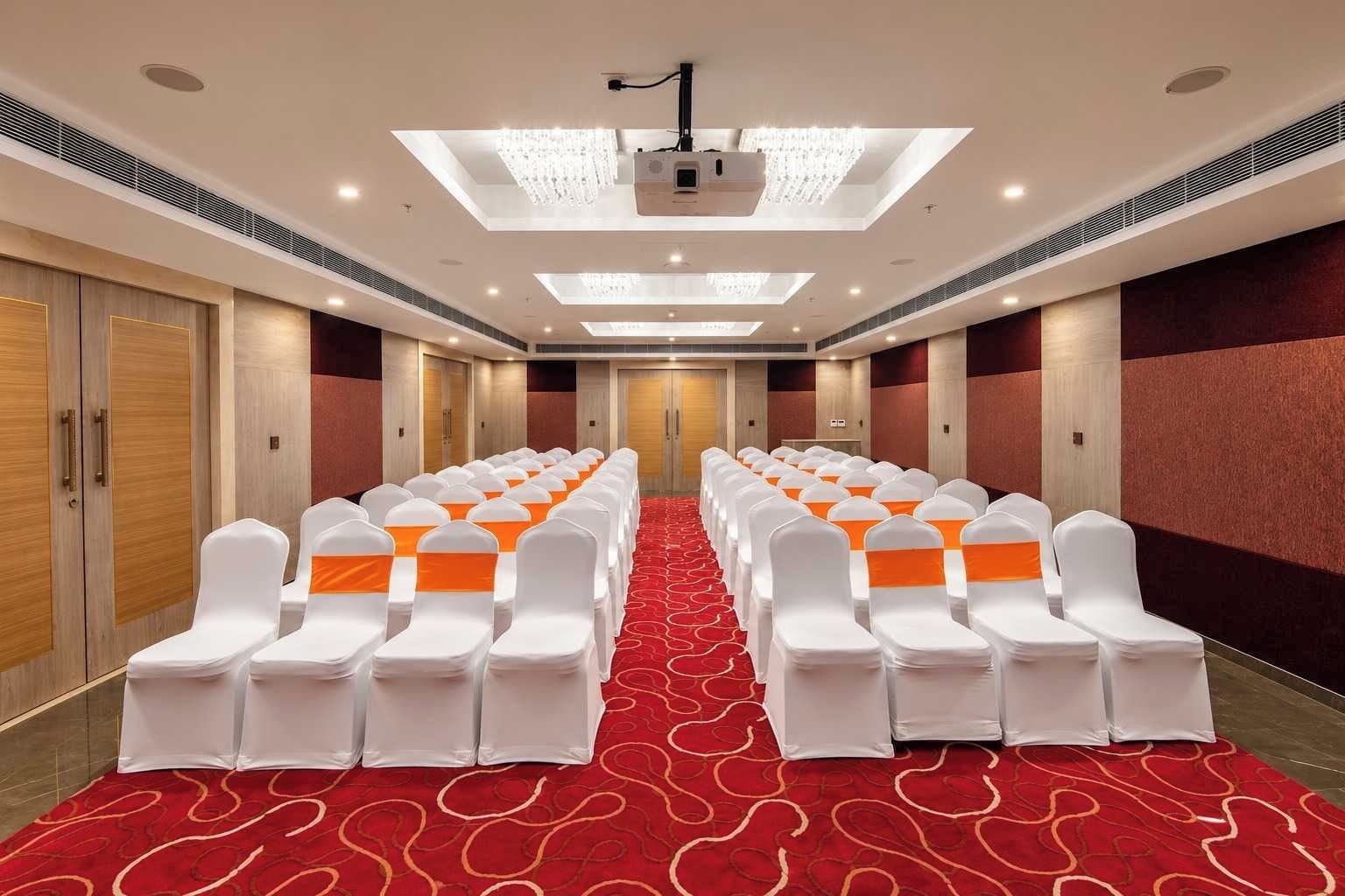 Uma sala de conferências de hotel com fileiras de cadeiras brancas com fitas laranja, um tapete com padrão vermelho e um projetor no teto.