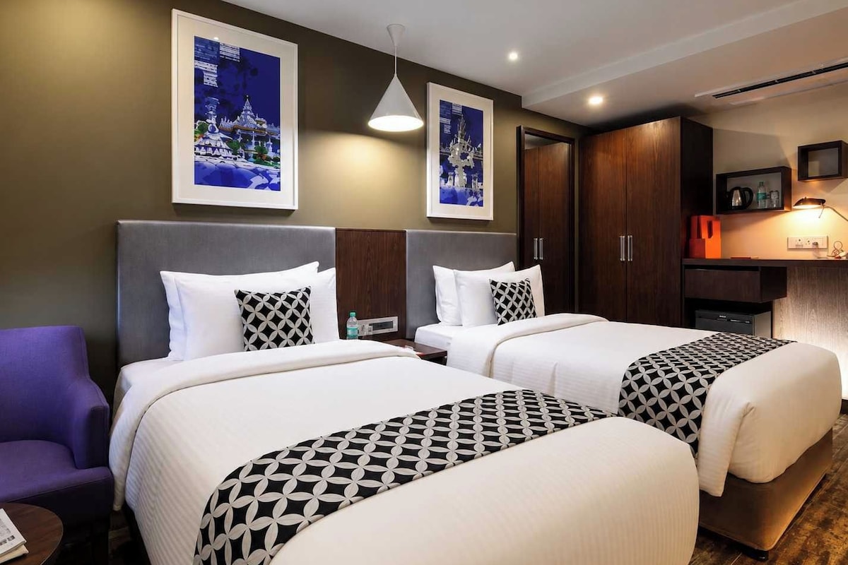 Ein Hotelzimmer mit zwei Betten, einem Schreibtisch, einem Stuhl und Kunstwerken an den Wänden.