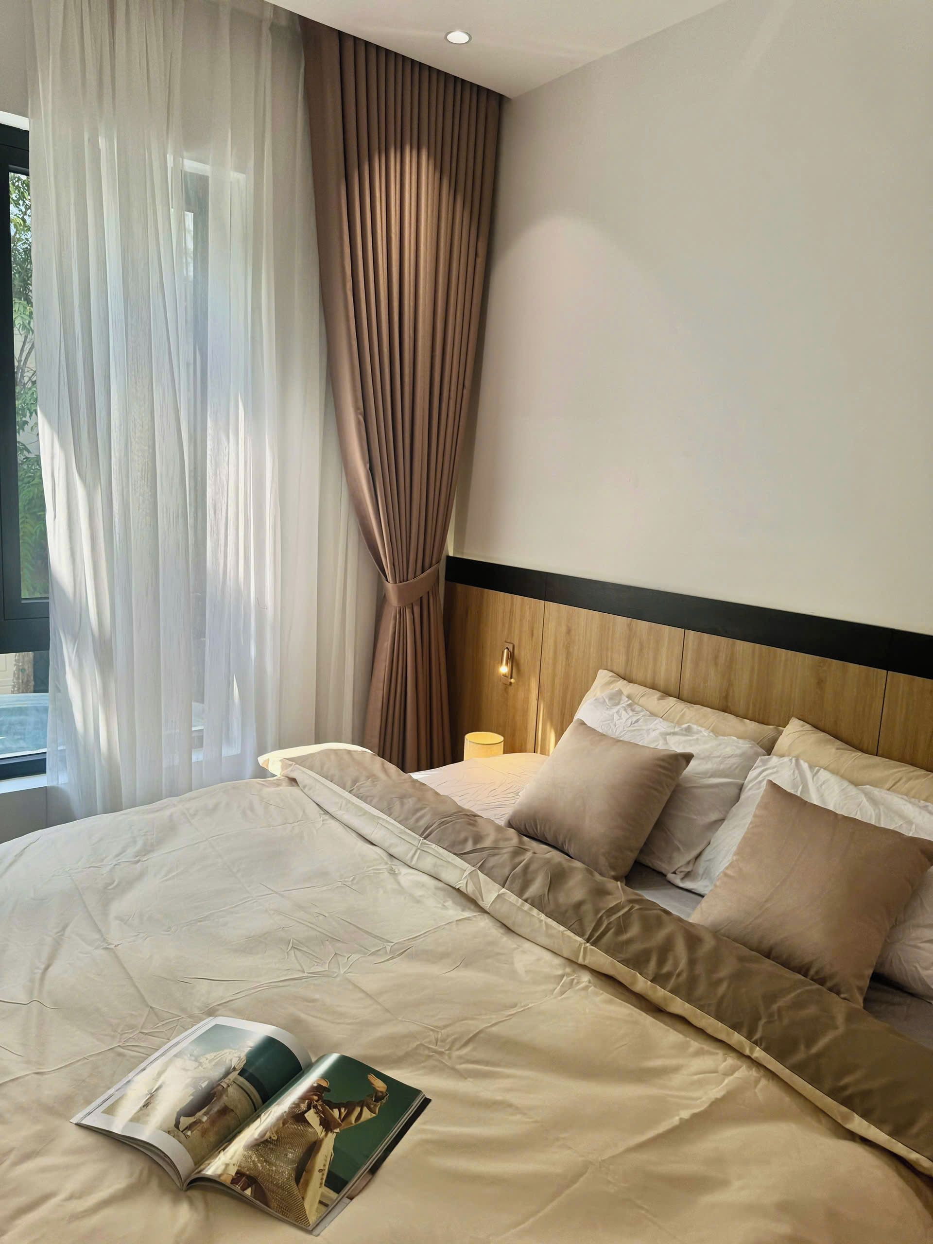 A neatly made bed with a magazine, a wooden headboard, and a window with sheer curtains.