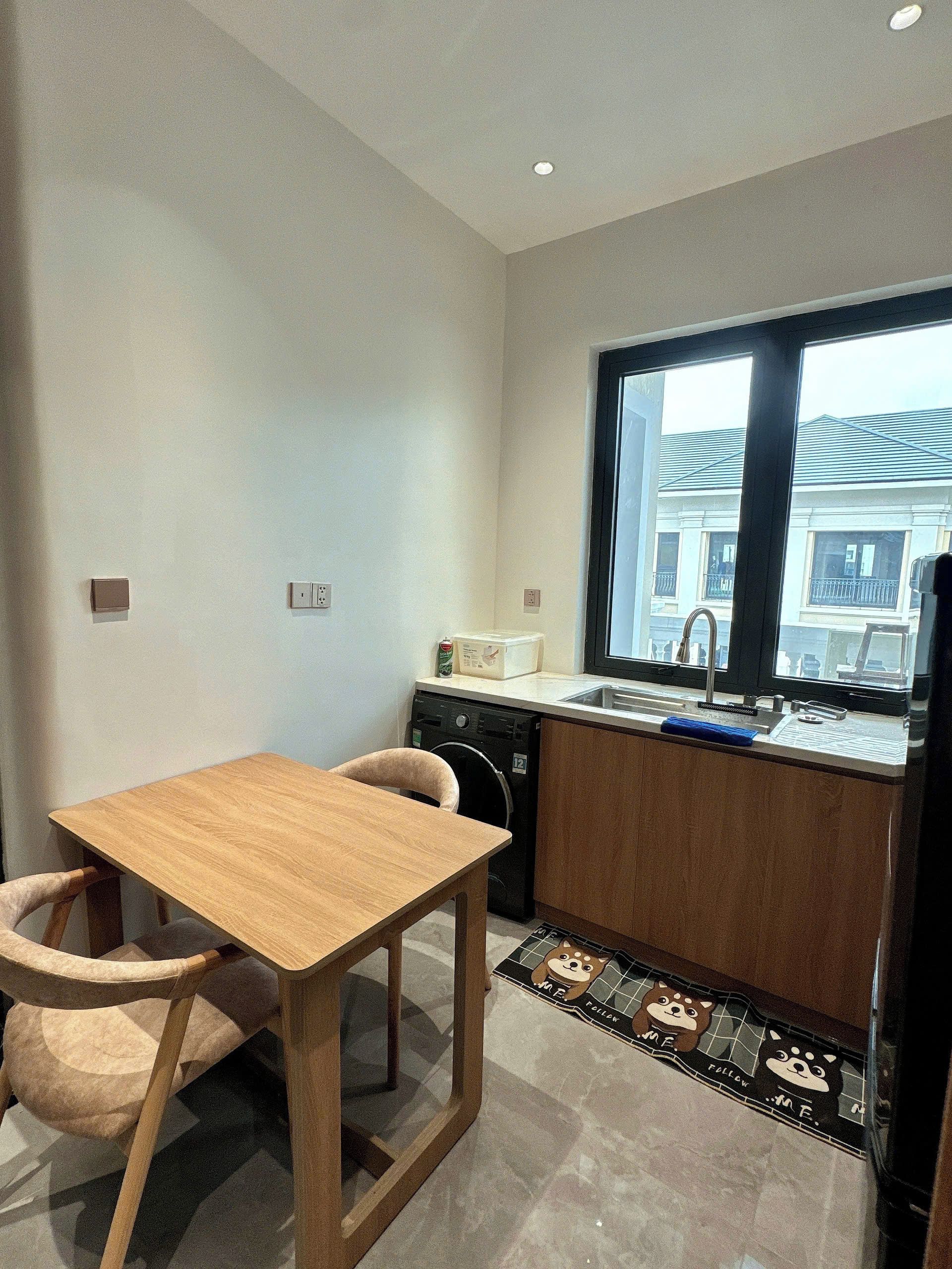A compact kitchen with a wooden table and chairs, a washing machine, and a window with a view of buildings.
