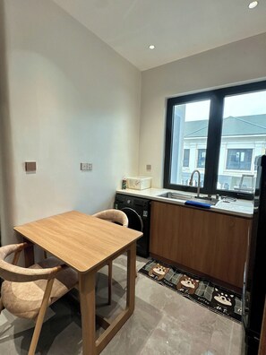 A compact kitchen with a wooden table and chairs, a washing machine, and a window with a view of buildings.