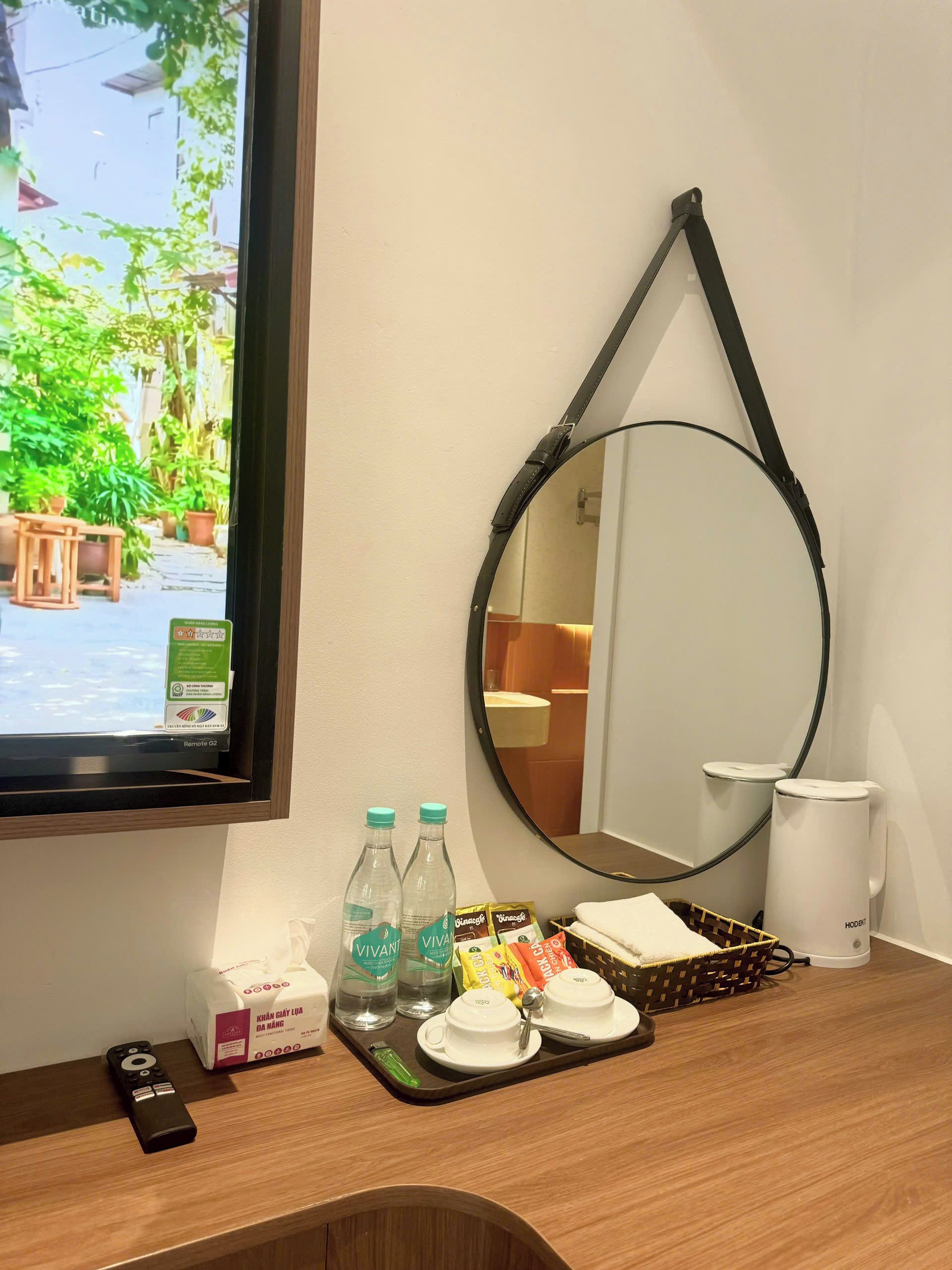 Ein Hotelzimmer mit einem Holztisch, einer Fernbedienung, einem Tablett mit Tassen und Snacks, einem Spiegel und einem Fenster mit Blick auf Grünflächen.