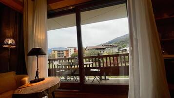 Une pièce avec un plancher en bois, une table avec une lampe, un canapé et une vue sur des immeubles et des montagnes, visible à travers une porte vitrée.