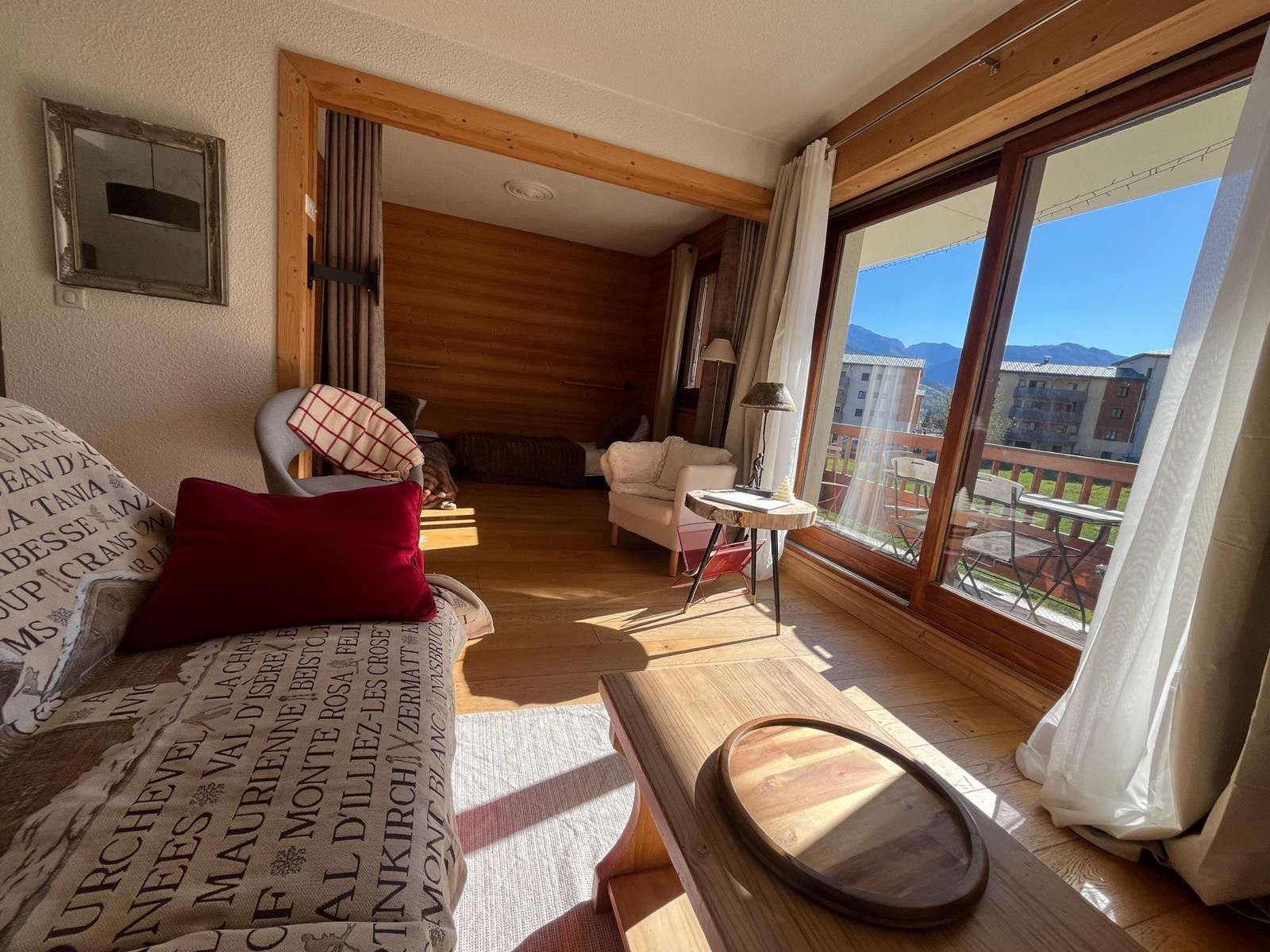 Una stanza accogliente con soffitto in legno, un divano con cuscini decorati, un tavolino e vista su un balcone con mobili da esterno.