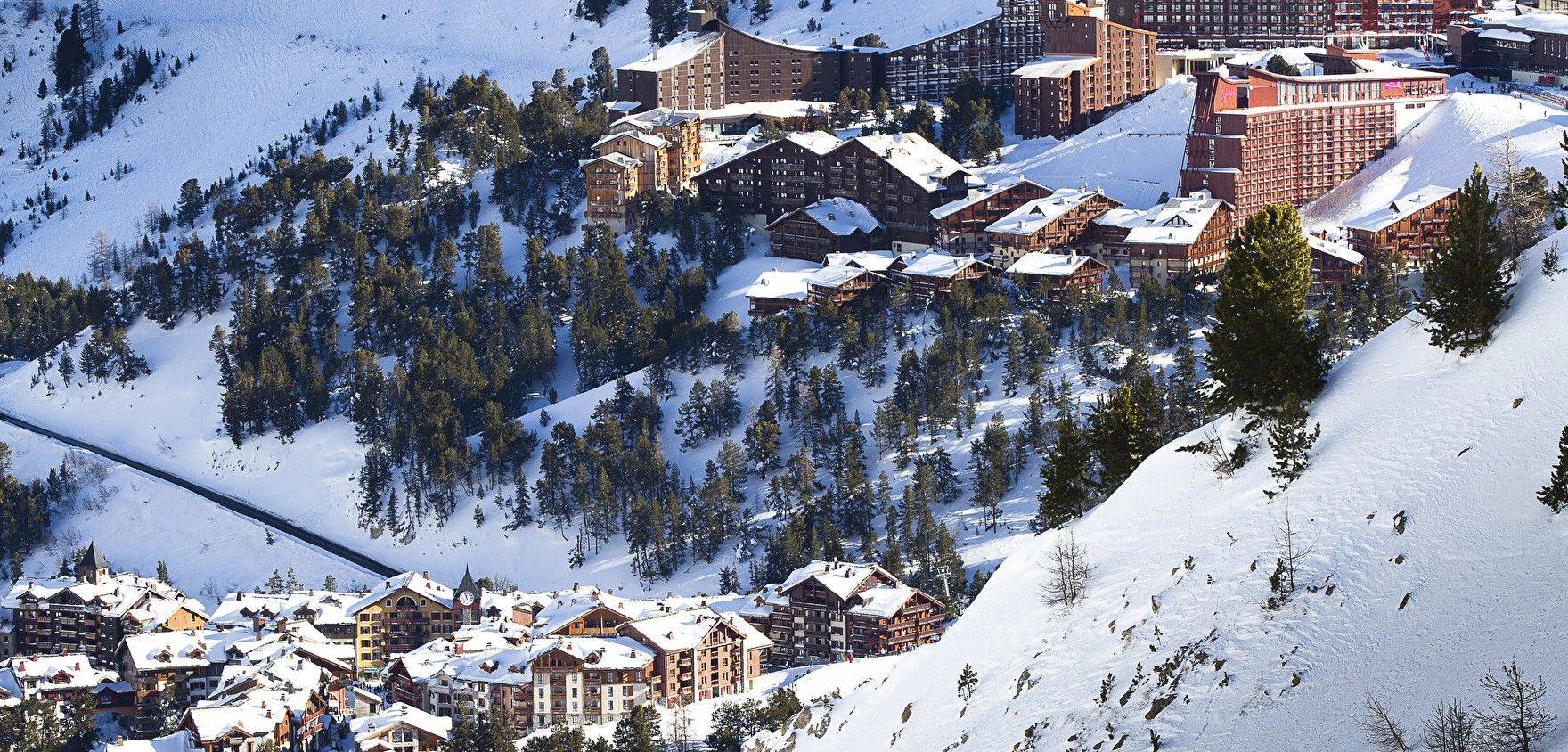 Ein schneebedecktes Gebirgendorf mit mehreren Gebäuden, darunter Hotels und Ferienhäusern, umgeben von Bäumen und Skipisten.
