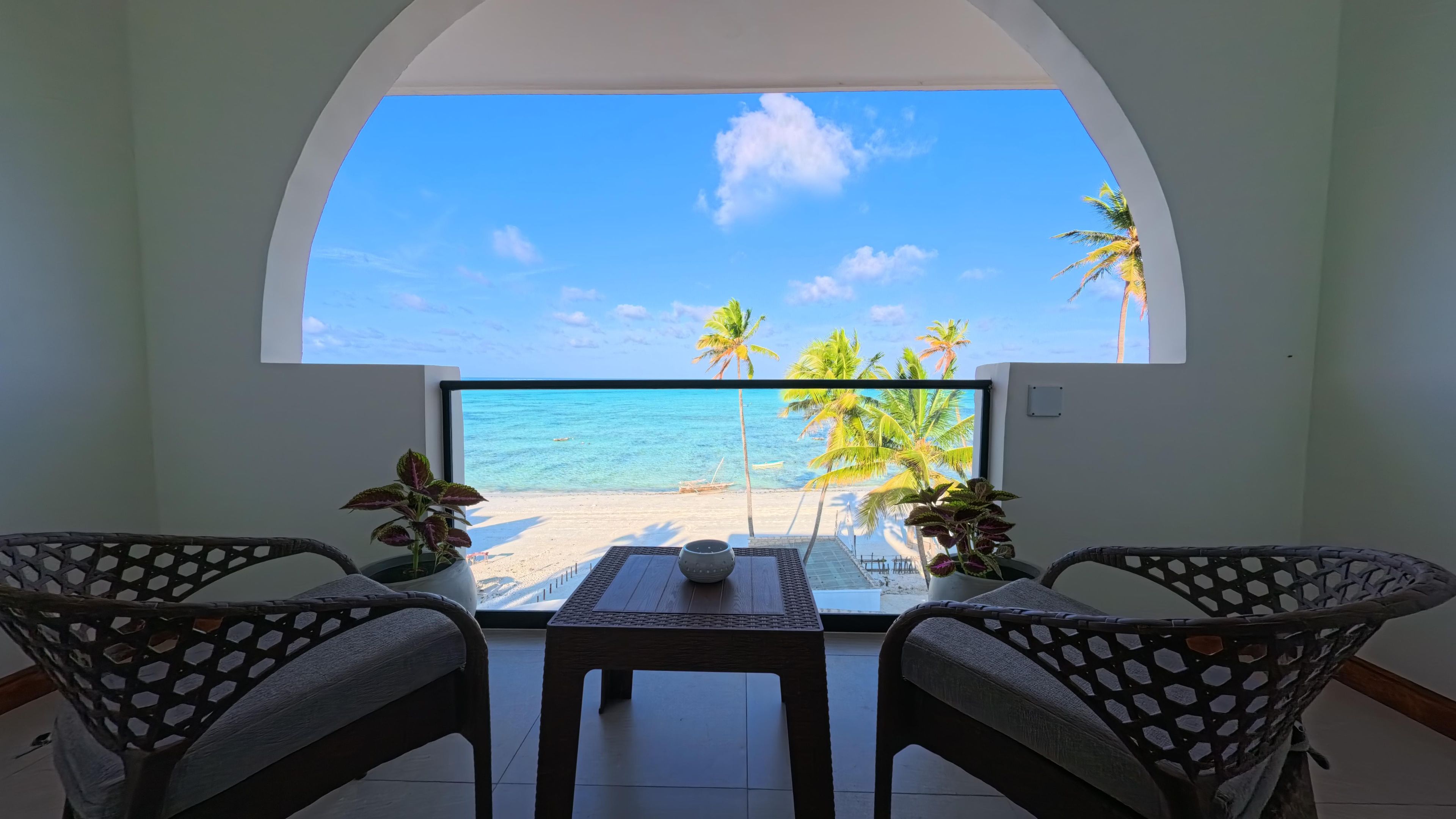 A room with a view of a beach, two wicker chairs, a table, and a potted plant.