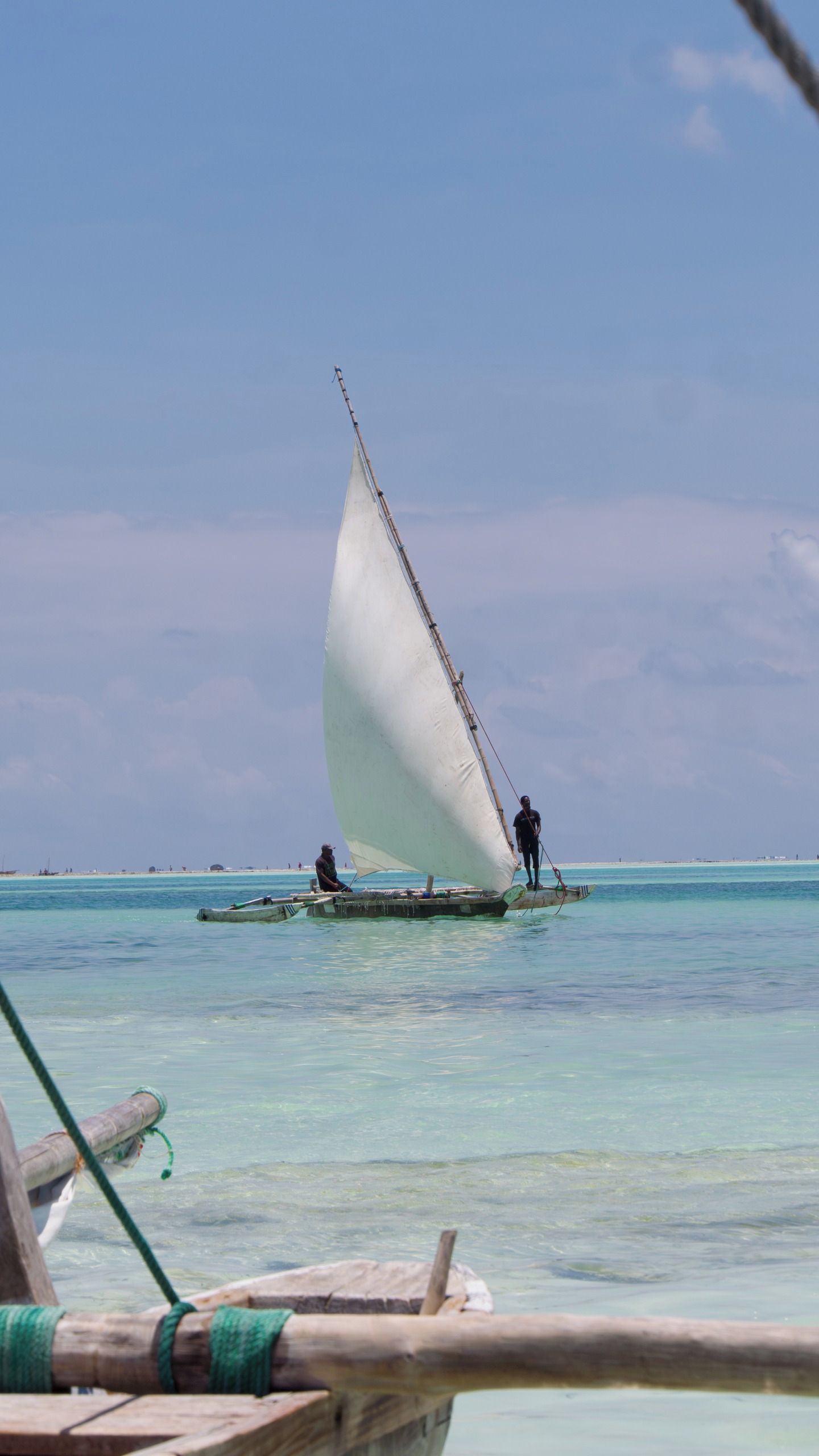 A traditional sailboat with a single sail, two people on board, and calm turquoise waters.