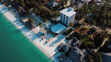 An aerial view of a coastal hotel with a beach, palm trees, and a large building with a flat roof.