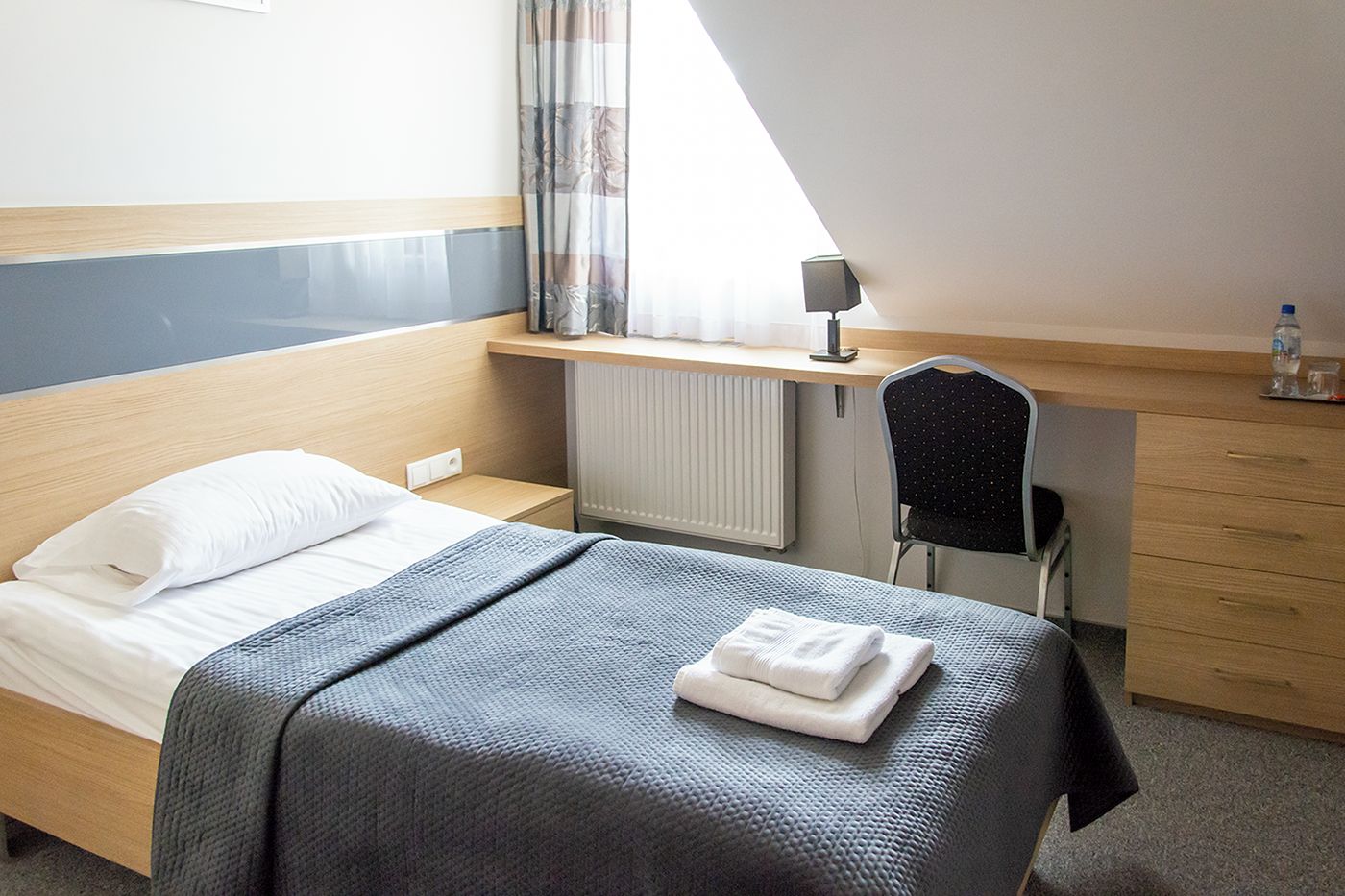 Una habitación de hotel con cama, escritorio, silla, radiador y ventana con cortinas.