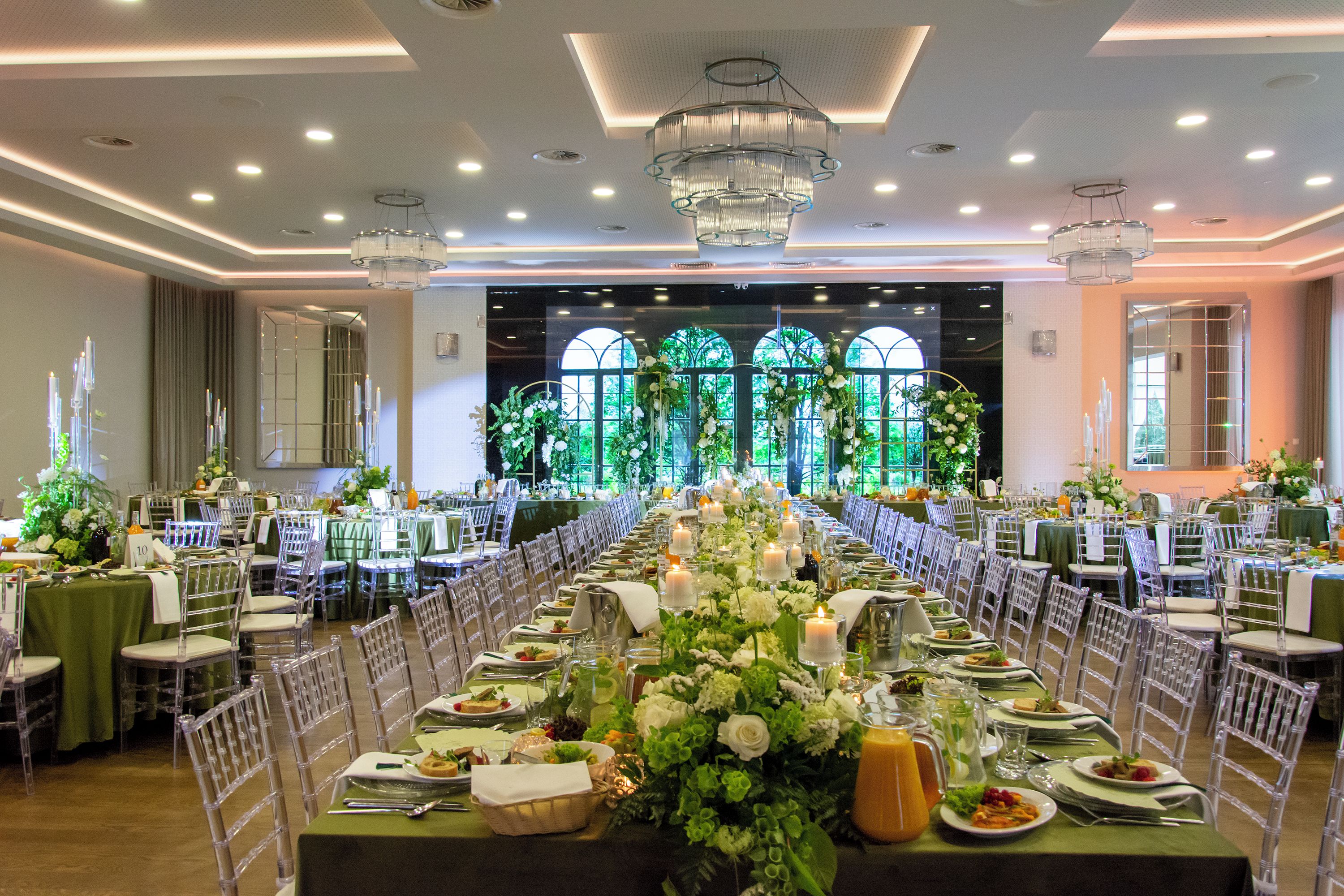 A banquet hall with long tables set for a formal event, featuring clear chairs, green tablecloths, and floral centerpieces.