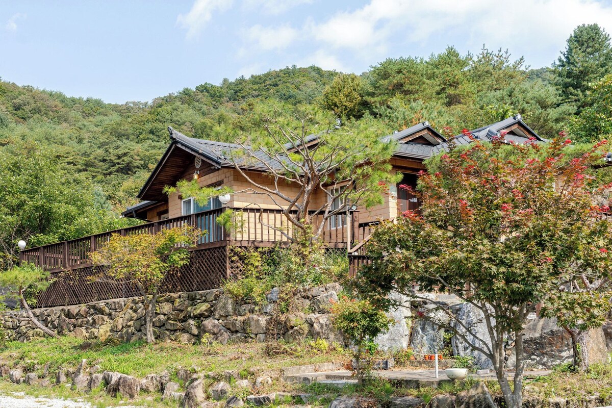 Uma casa tradicional em estilo japonês, fachada de madeira, parede de pedra e vista para uma encosta arborizada.