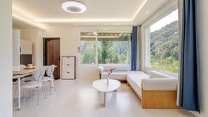 Uma sala de estar moderna com área de jantar, sofá, mesa de centro e vista para uma encosta arborizada.