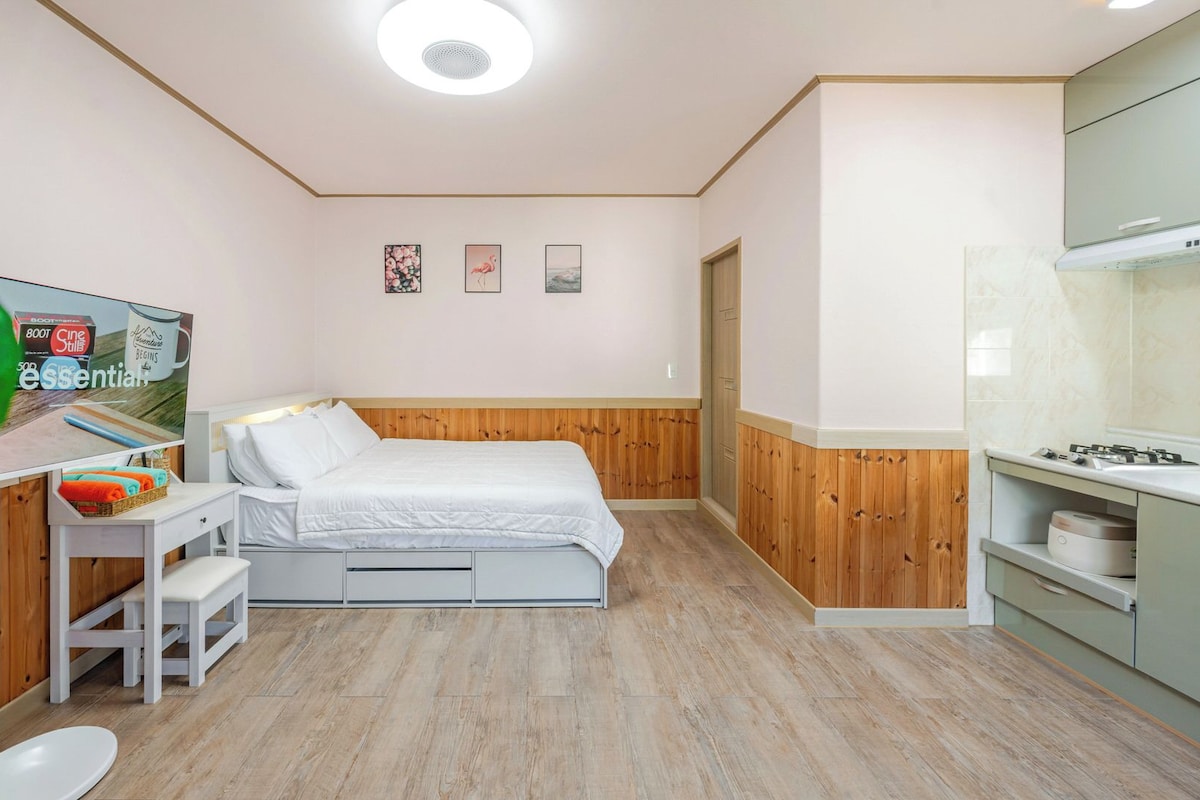 Quarto de hotel compacto com cama, mesa de cabeceira e pequeno espaço de cozinha.