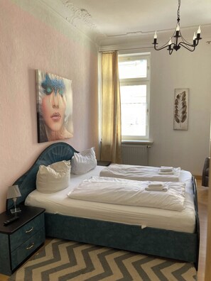 Ein Hotelzimmer mit Bett, Nachttisch, Lampe, einem Gemälde, einem Fenster mit Vorhängen und einer Kristallleuchtre.