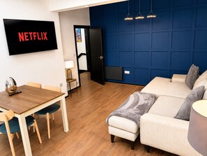 A modern living room with a flat-screen TV displaying 'NETFLIX,' a dining area with a wooden table and chairs, and a blue accent wall. - Modern 2 Bed Flat in Heart of Warrington City (Warrington)
