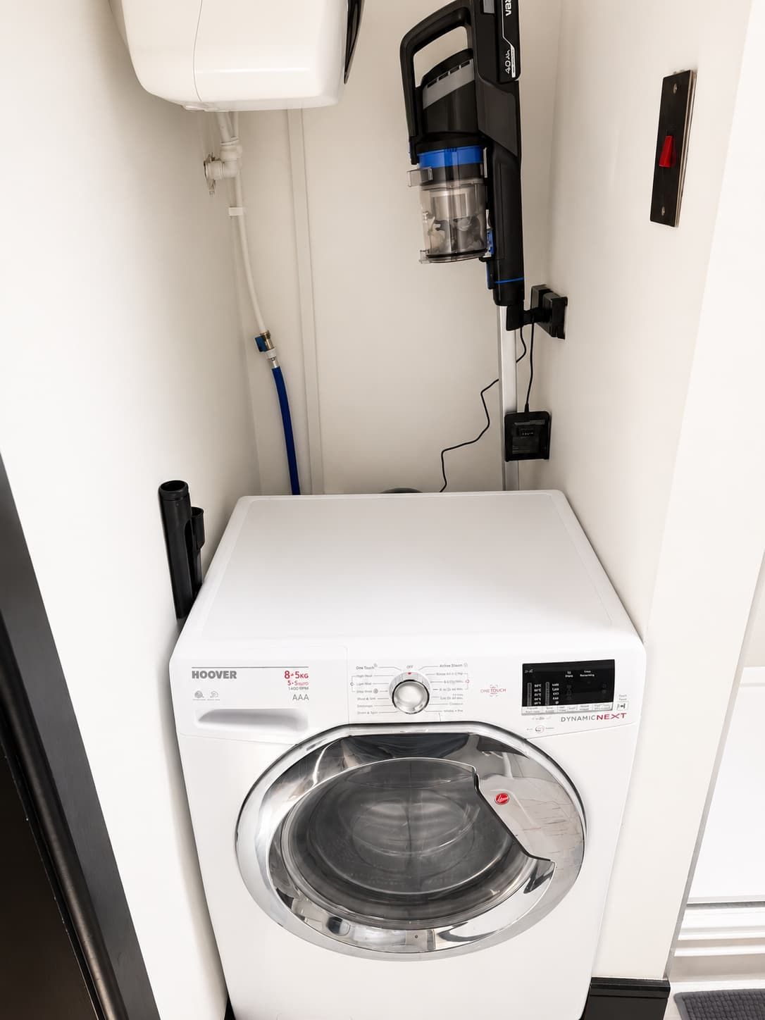 A compact, white, front-loading washing machine with a digital control panel, a hose, and a power outlet on the wall.