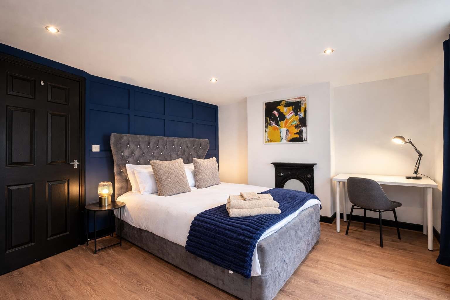Une chambre à coucher comprenant un lit, une table de chevet, un bureau, une chaise et une œuvre d’art accrochée au mur.