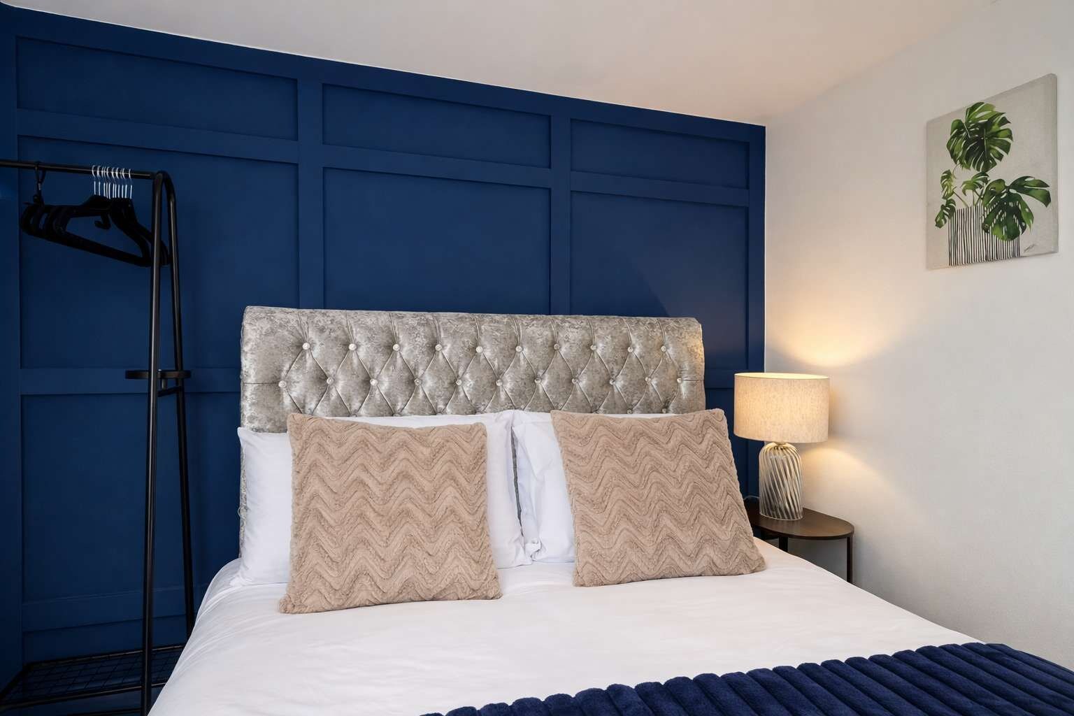 Een slaapkamer met een gestuukt hoofdeinde, twee kussens, een bedlamp en een wandplant.