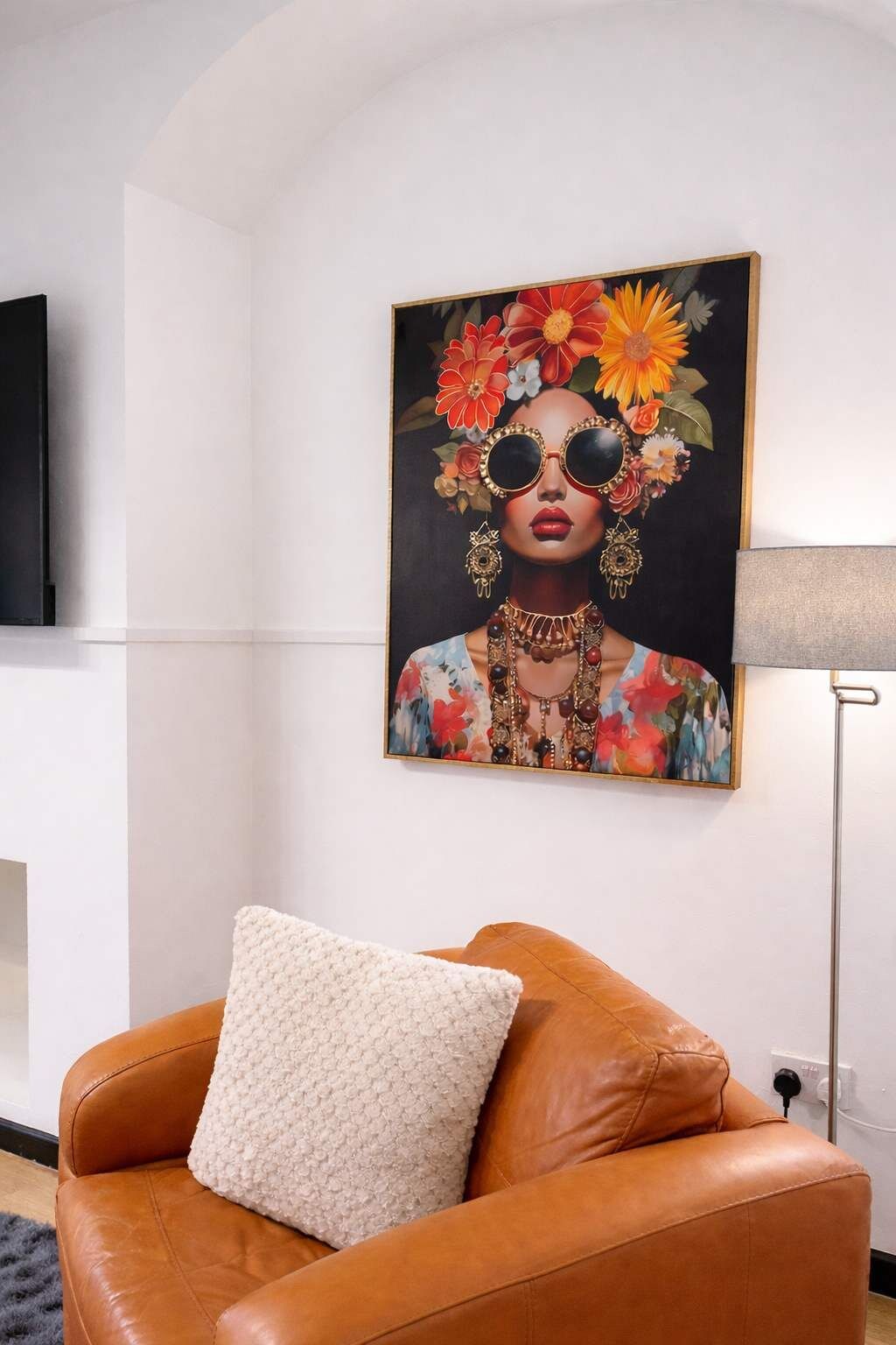 Een kamer met een bruine leren bank, een wit kussen met structuur en een schilderij met een bloemenmotief aan de muur.