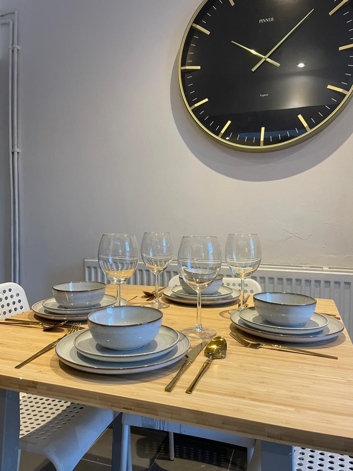 Une table à manger dressée avec des assiettes, des bols, des verres à vin, des couverts et une horloge murale.