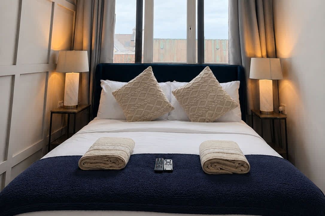 A hotel room with a bed, two pillows, two bedside lamps, and a remote control on the bed.