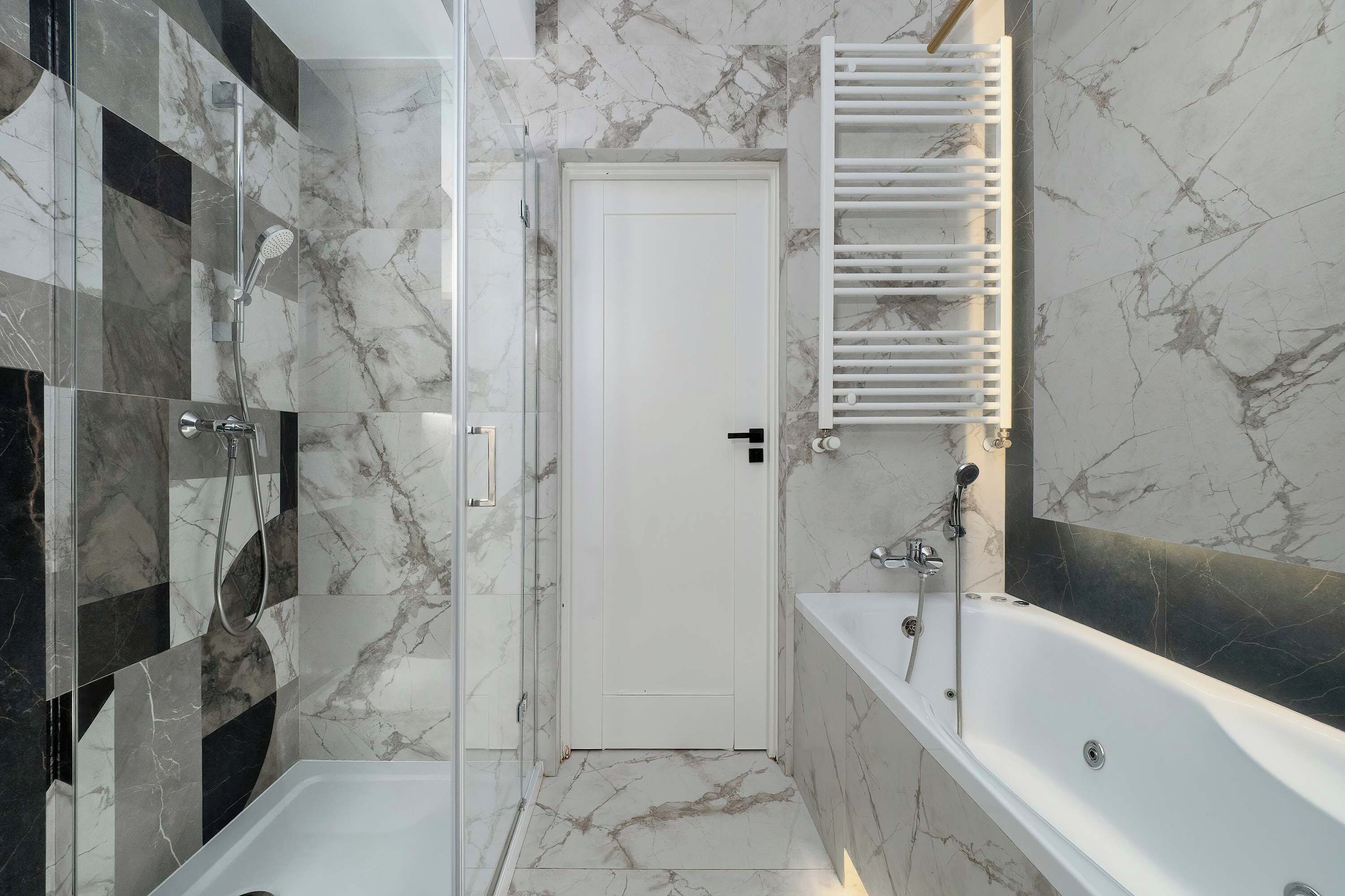 Large marble-patterned tiles cover the walls of this sleek bathroom, which includes a modern white bathtub. A white door and a heated towel rack are positioned at the far end of the room.