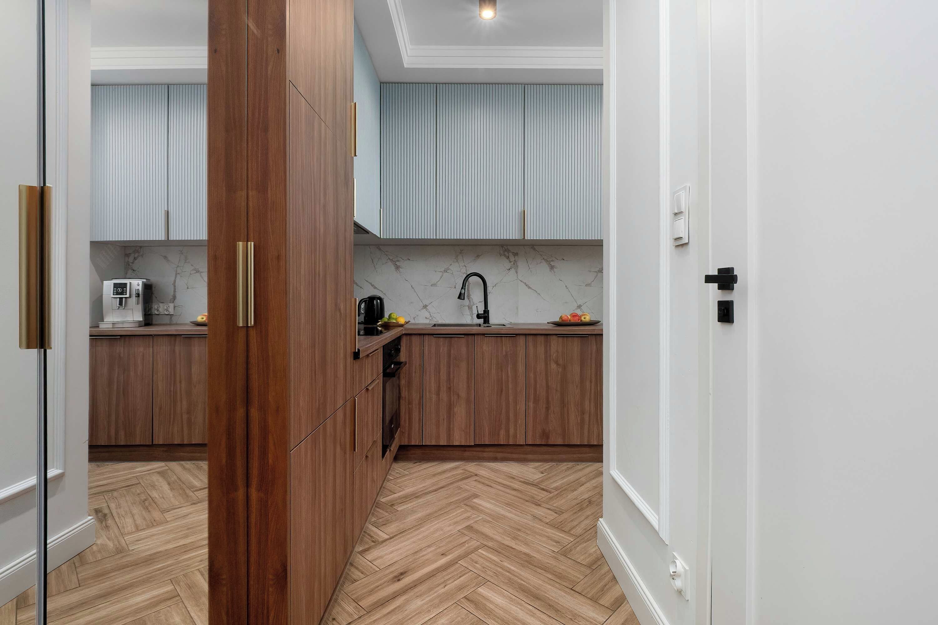 A hallway view looks directly into a modern kitchen area. The warm wood tones of the lower cabinets and the light herringbone-patterned floor create a welcoming entryway.