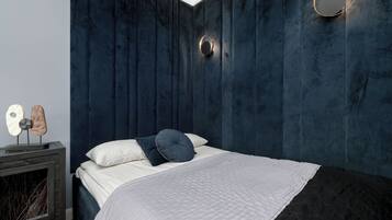 Quarto com parede azul escura, cama com roupa de cama branca e azul escura, espelho redondo e uma pequena escultura em uma mesinha de cabeceira.