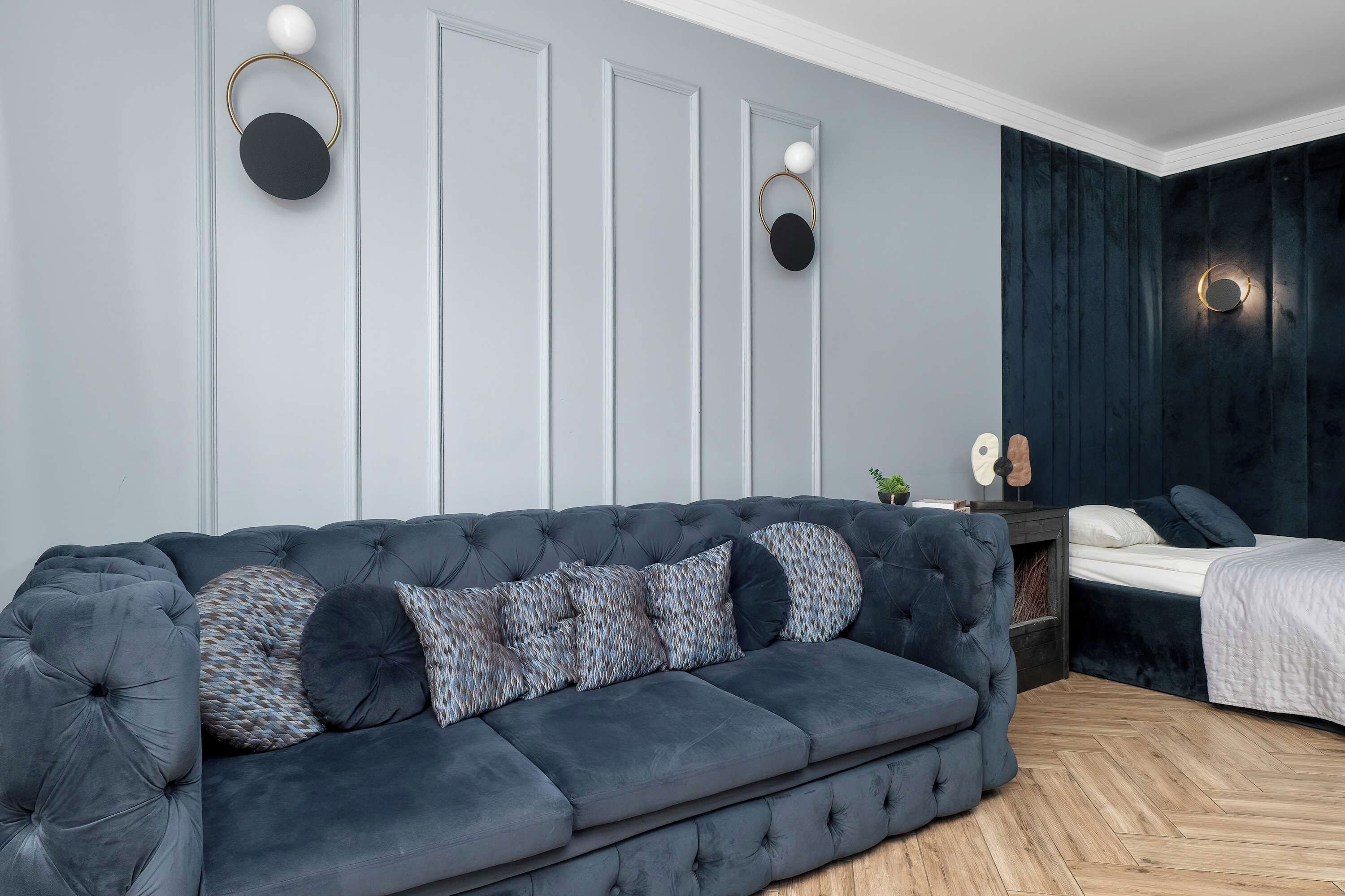 A modern bedroom with a dark blue upholstered sofa, a bedside table with a lamp, and a bed with a white duvet.