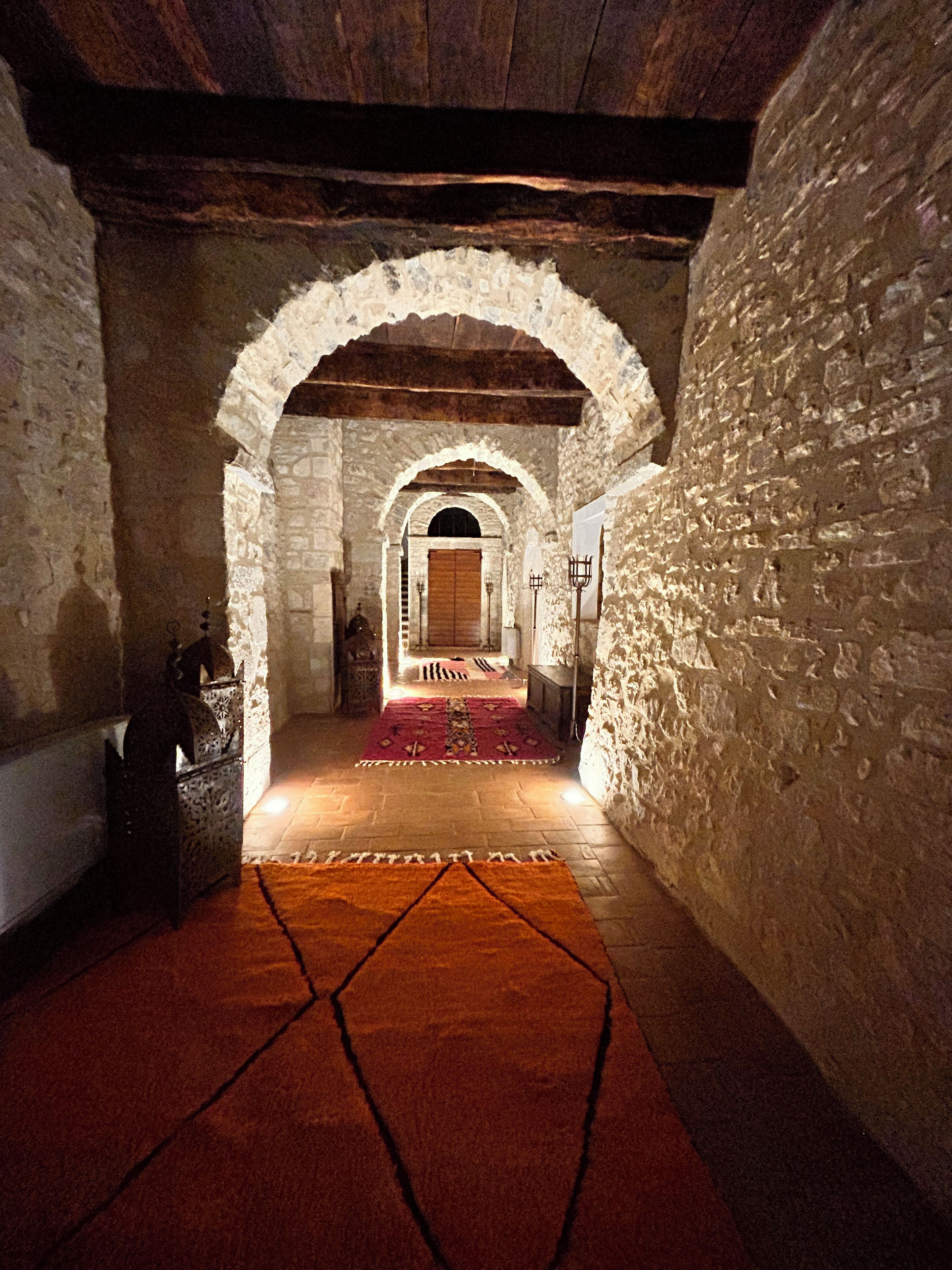 A corridor with stone walls and wooden beams, leading to a room with a patterned carpet.