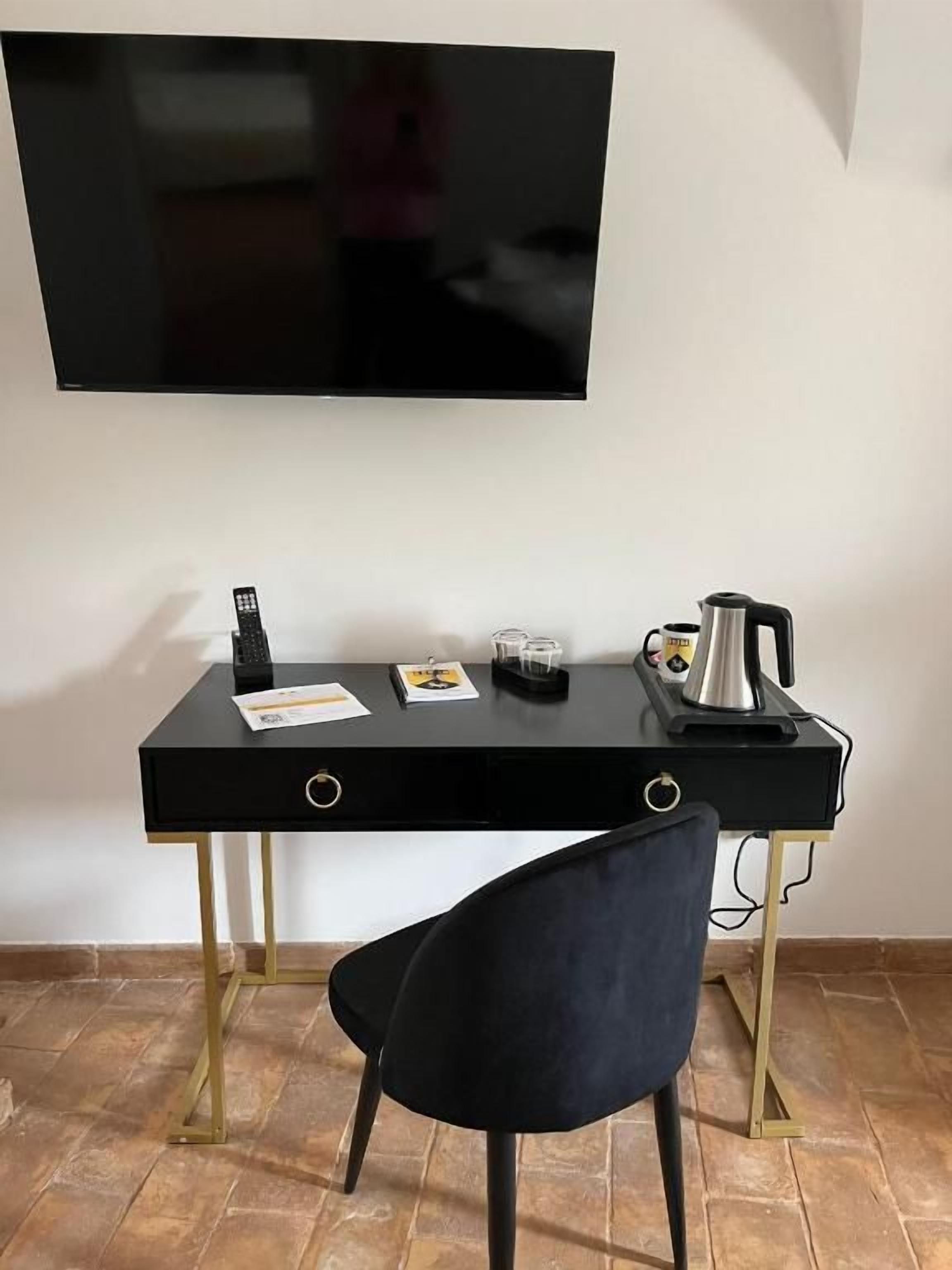 Una habitación con un escritorio negro, una silla, un televisor de pantalla plana montado en la pared y una cafetera sobre el escritorio.