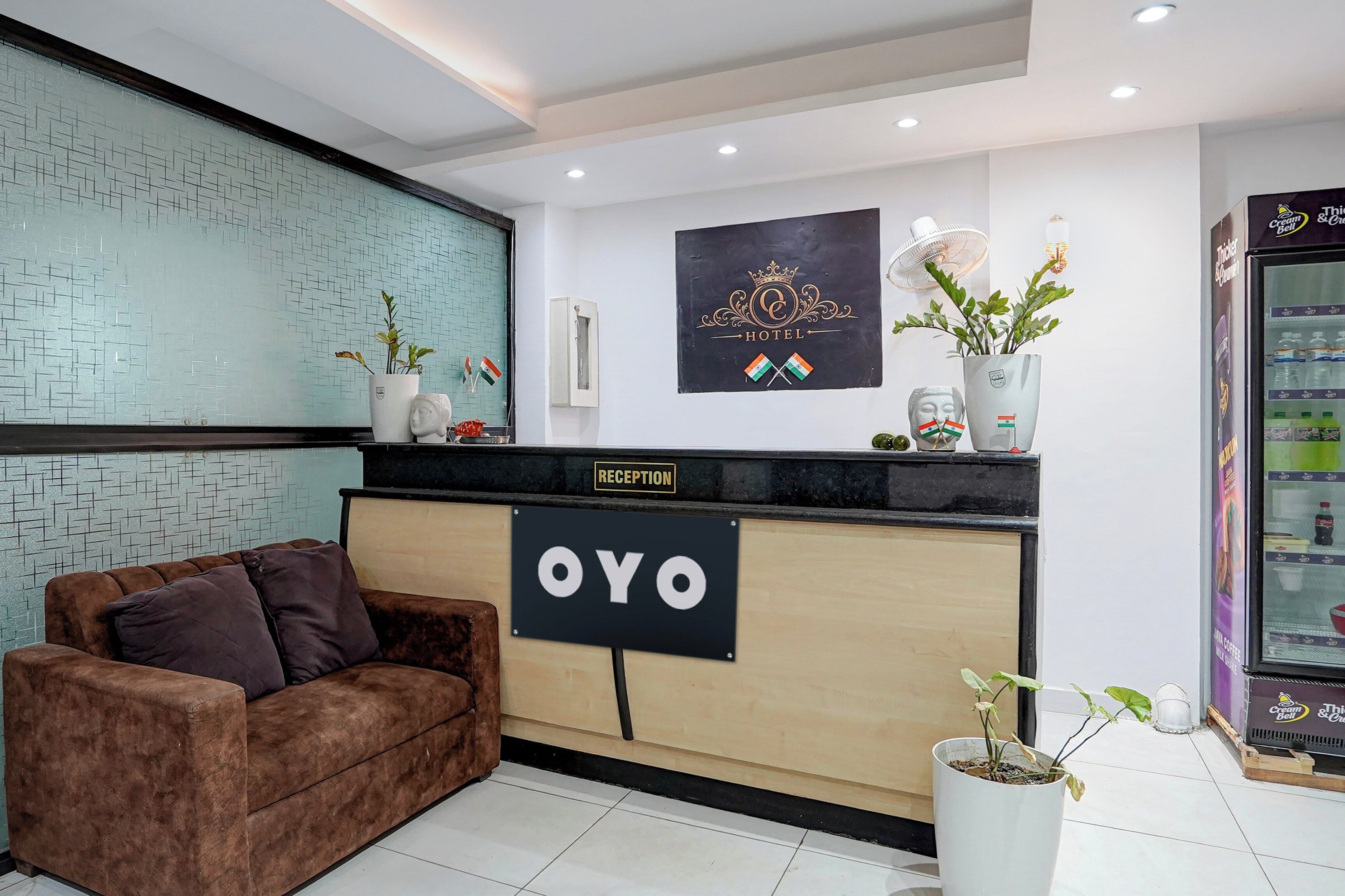 Uma área de recepção com balcão identificado como 'OYO', um sofá, um ventilador fixado na parede e uma geladeira abastecida com bebidas.